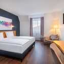 Modernes Hotelzimmer mit Doppelbett, farbenfrohem Kunstwerk an der Wand, gemütlicher Sitzecke und großem Fenster.