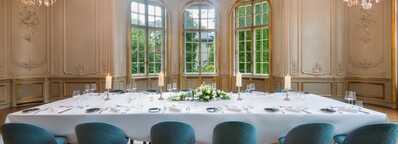 A luxurious dining room features a long, elegantly set table with blue chairs and ornate chandeliers illuminating the space.