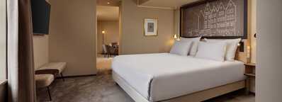A modern bedroom features a large bed with white linens, a wall-mounted TV, and an adjoining seating area in a stylish hotel.