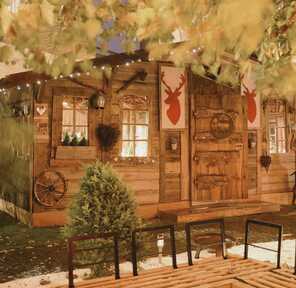 A cozy wooden cabin decorated with deer motifs is illuminated warmly, surrounded by lush foliage and rustic decor.