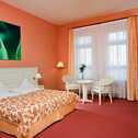 Gemütliches Zimmer mit einem großen Bett, einem kleinen Tisch mit zwei Stühlen und einem beruhigenden grünen Kunstwerk an der Wand.