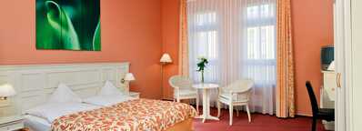 A cozy hotel room features a double bed with floral bedding, a small seating area, and a large window with sheer curtains.