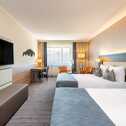 A modern hotel room features two neatly made beds, a flat-screen TV, and a seating area by large windows.