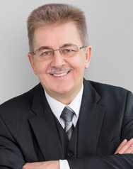 A smiling man in a suit crosses his arms confidently against a neutral background, embodying leadership and professionalism.