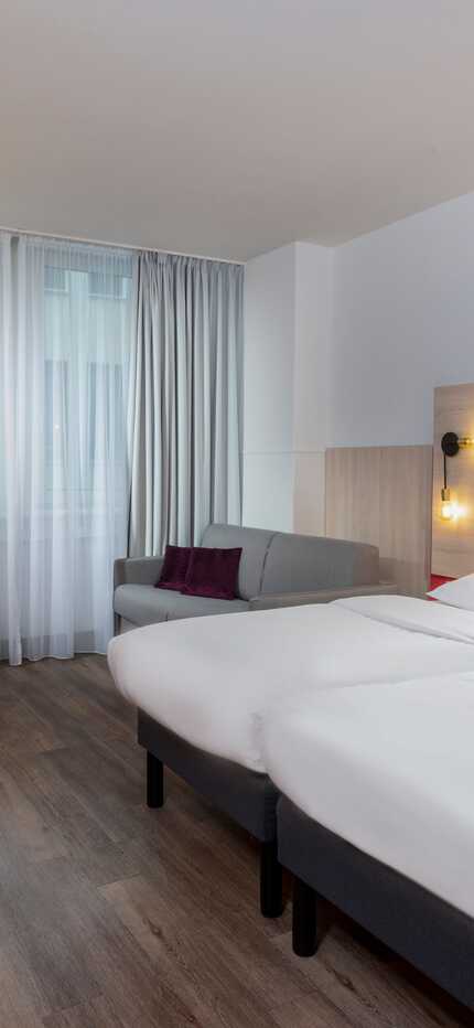 Helles Hotelzimmer mit zwei Betten, einem grauen Sofa und gelbem Stuhl bietet Komfort für Familienaufenthalte.