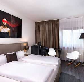 A modern double room features a large bed, stylish artwork, and a cozy seating area by the window.