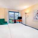 A bright hotel room features a large bed, modern art, a green chair, and a window with a city view.