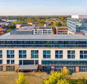 A modern, multi-story building with large windows stands amidst a scenic urban landscape under a clear blue sky.