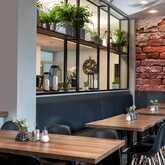 Modernes Restaurant mit Holztischen, schwarzen Stühlen und dekorativen Pflanzen vor einer Backsteinwand.