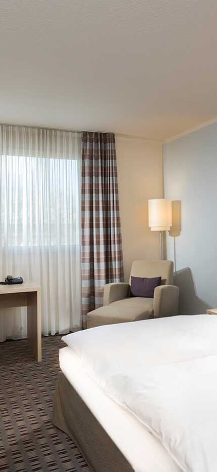 A standard hotel room features a cozy bed, a TV, and a desk by the window with sheer curtains.