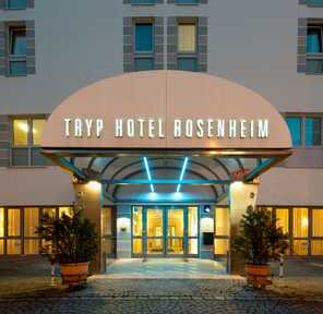 The illuminated entrance of Tryp Hotel Rosenheim glows warmly at dusk, flanked by potted plants and large windows.