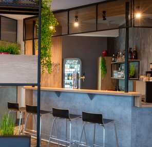 Eine moderne Bar mit Holztresen, Barhockern und Pflanzen, neben einem gut bestückten Kühlschrank und einer Kaffeemaschine.