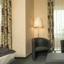 A cozy double room features two armchairs, a glass table, and elegant floral curtains beside a comfortable bed.
