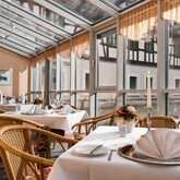 A cozy restaurant with wicker chairs and elegantly set tables overlooks a scenic courtyard through large glass windows.