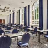 A sunlit conference room with blue chairs and long tables is set up for a seminar, featuring large windows and curtains.