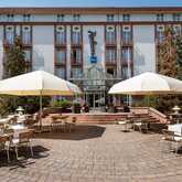 The exterior of a hotel features a charming patio with tables, chairs, and large umbrellas under a clear blue sky.