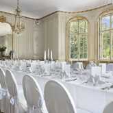 Ein elegant gedeckter Tisch mit weißen Stühlen und Kerzenleuchtern steht in einem prunkvollen Musikzimmer vor großen Fenstern.