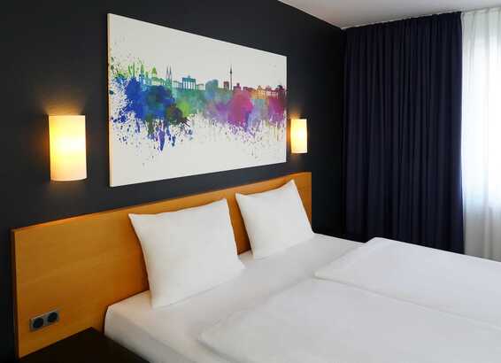 A modern hotel room features a double bed, vibrant cityscape artwork, and soft lighting for a cozy ambiance.