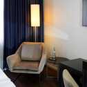 A modern hotel room features a cozy armchair, a tall lamp, and a small table with a water bottle and glasses for a superior experience.