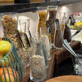 A breakfast buffet features a variety of cereals and grains in glass jars, with fresh fruit in a basket nearby.
