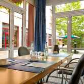 A bright meeting room with a conference table set with blue folders and water, overlooking a leafy courtyard through large windows.