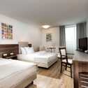 A modern business hotel room with twin beds, a desk, and a TV, featuring minimalist decor and natural light through a window.