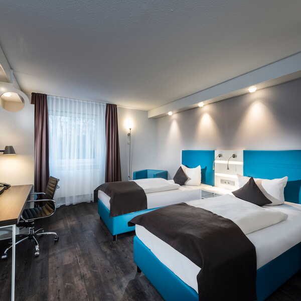 A bright twin bedroom features modern decor, two beds, a work desk, and large windows.