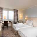 A well-lit standard twin bedroom features two neatly made beds, a desk with a chair, and a flat-screen TV.