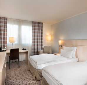 A well-lit standard twin bedroom features two neatly made beds, a desk with a chair, and a flat-screen TV.