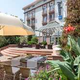 The hotel exterior features a sunlit terrace with umbrellas, dining tables, and lush greenery for a relaxing outdoor experience.
