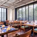 Elegant restaurant interior with neatly set tables, wooden chairs, and large windows with blinds overlooking greenery.