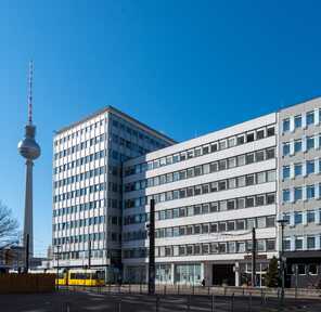 The exterior of a modern building in Berlin is shown against a clear blue sky, with the TV tower visible in the background.