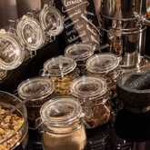 Nahaufnahme einer Auswahl an Müslizutaten und Toppings, präsentiert in Glasbehältern und Weckgläsern. Erkennbar sind verschiedene Kerne, Nüsse oder Trockenfrüchte, die zum Verfeinern von Müsli oder Joghurt dienen. Ein Mörser und weitere Behälter sind im H