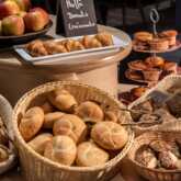A close-up of a lavish breakfast buffet with several baskets full of different rolls and types of bread in the foreground. Behind it is a platter of croissants, a tiered stand with muffins and donuts and a bowl of fresh red apples. A small sign with the w