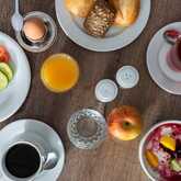 A top view of a laid breakfast table with various foods and drinks. You can see a plate with cold cuts (sausage and ham) and vegetables (tomato, cucumber), a boiled egg, bread rolls, a glass of orange juice, a glass of water, an apple, a cup of coffee, a