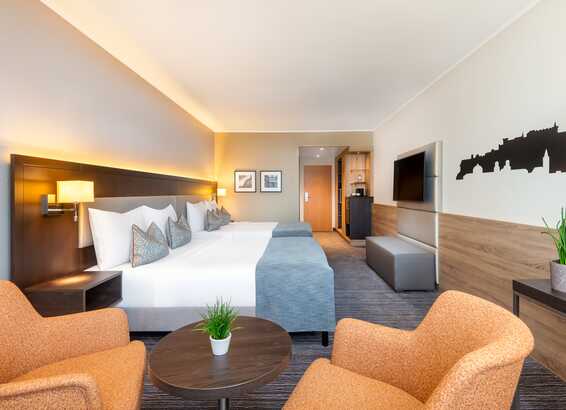 A modern hotel room features a neatly made bed, cozy chairs, and a city skyline wall design.