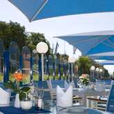 A modern outdoor terrace features blue chairs, white table settings, and flowers under large blue umbrellas on a sunny day.
