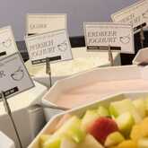 A breakfast buffet display features assorted yogurts and muesli with labels, alongside a bowl of fresh fruit.
