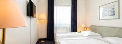 A bright twin bed room features modern decor, a wall-mounted TV, and ample natural light from a large window.
