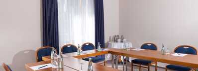 A well-lit meeting room with arranged tables, chairs, notepads, and bottled water awaits attendees.