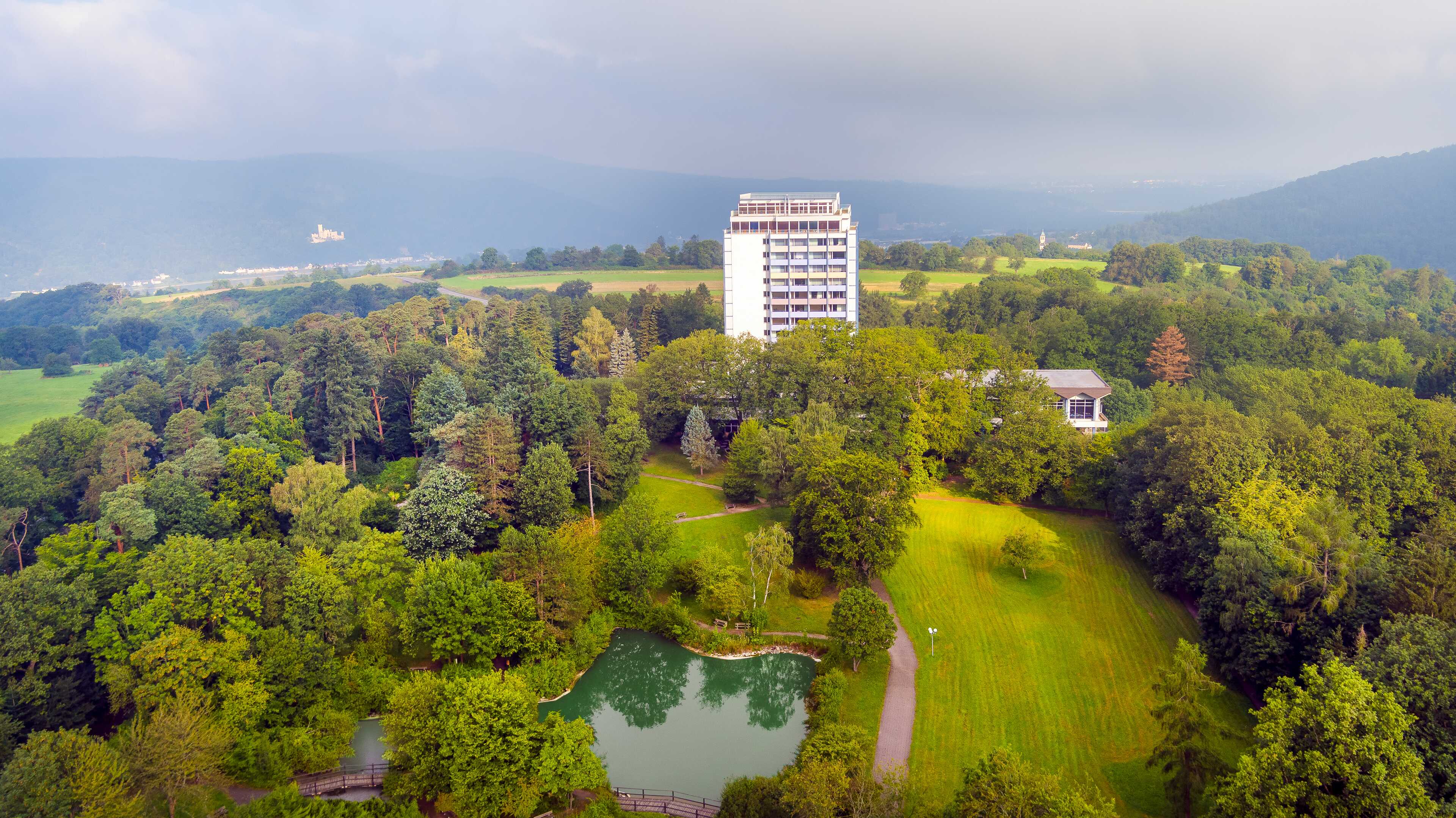Aerial view of a tall building surrounded by lush greenery and a small pond, set against distant hills.