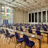 A spacious classroom with rows of blue chairs and tables is set for a lecture, with natural light streaming through large windows.