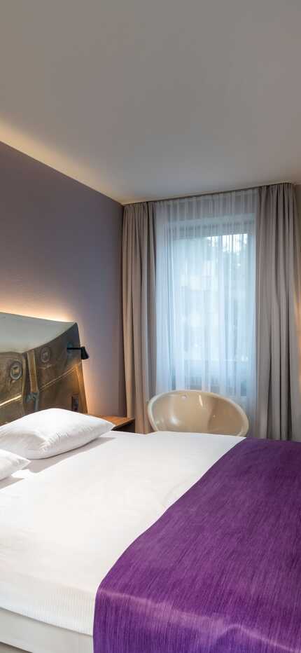 A stylish double room features a bed with a unique headboard, a purple throw, a desk with a chair, and a wall-mounted TV.