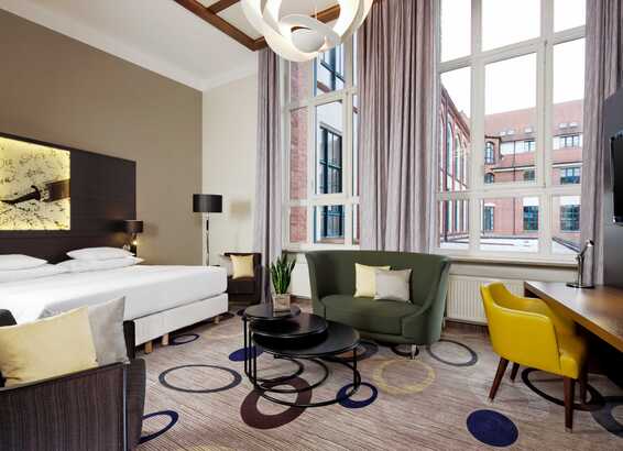 A modern hotel room features a cozy seating area, large windows with city views, and a stylish bed with soft lighting.