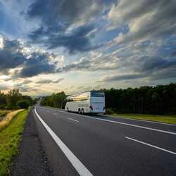 A white tour bus travels on a scenic highway at sunset, with dramatic clouds and lush greenery lining the road.