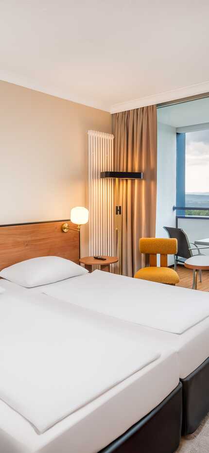 A modern hotel room with twin beds and a balcony overlooking a scenic landscape, featuring stylish decor and warm lighting.