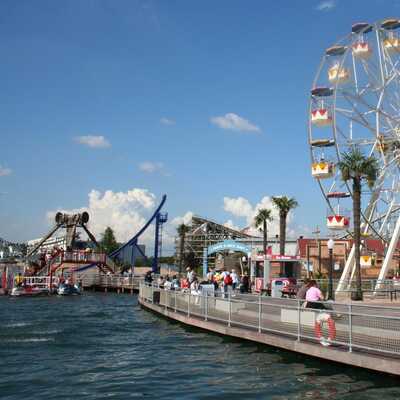 Ein Riesenrad dreht sich im sonnigen Moviepark Bottrop, umgeben von Palmen und fröhlichen Besuchern entlang des Wassers.