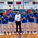 A group of seven men in blue shirts and beige pants pose confidently in a sports arena.