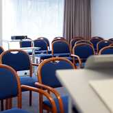 A seminar room with rows of empty blue chairs and a projector cart awaits attendees.