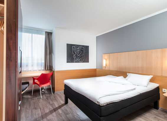 A modern single room features a cozy bed, a bright red chair by the window, and a stylish wall art piece.